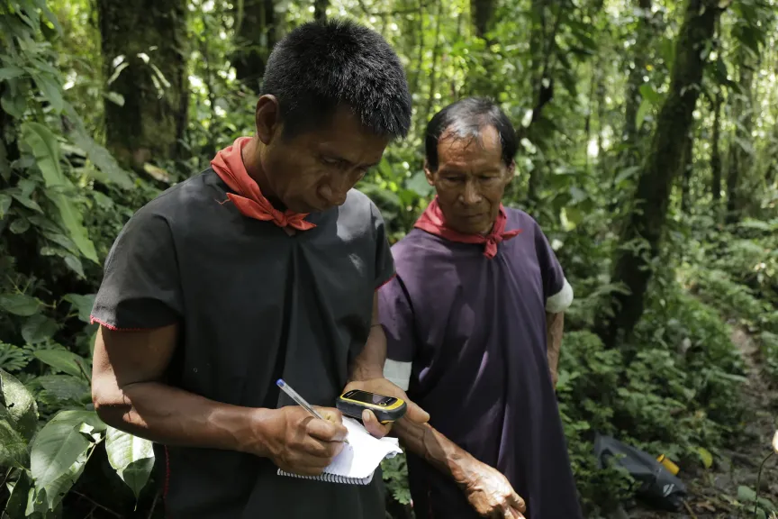 Two community members in the forest, one recording field notes while holding a GPS unit.