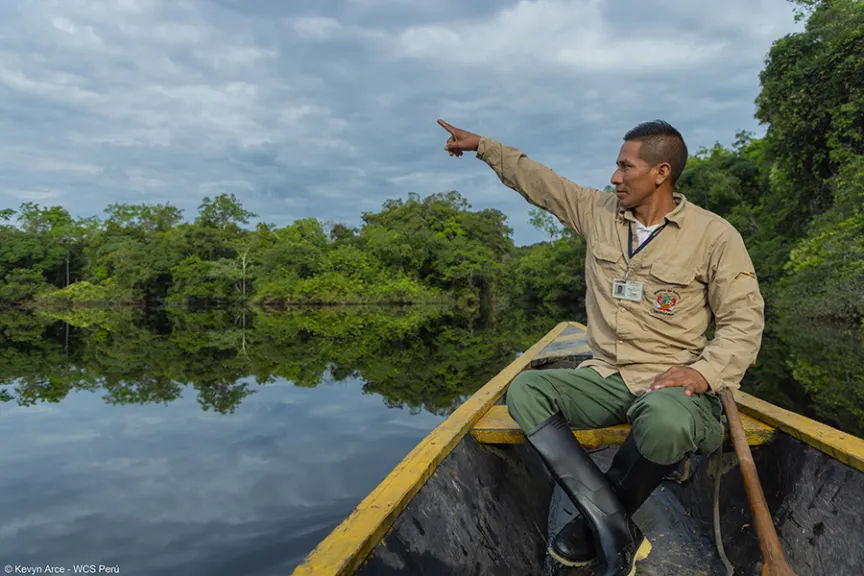 A park ranger in a wooden canoe points across a still river lined with forest.